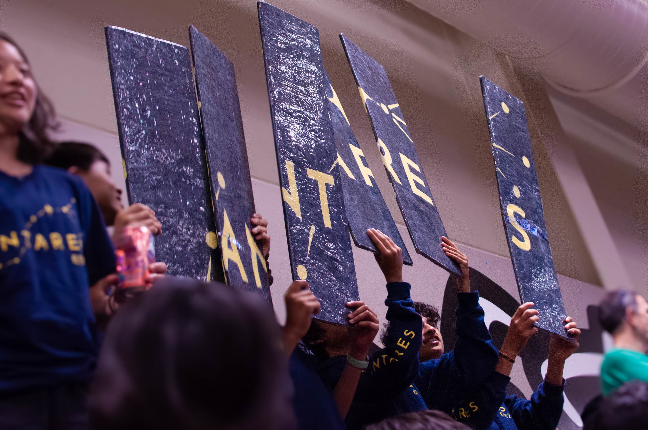 Antares students in the stands at a competition, sponsor logos on the backs of their shirts